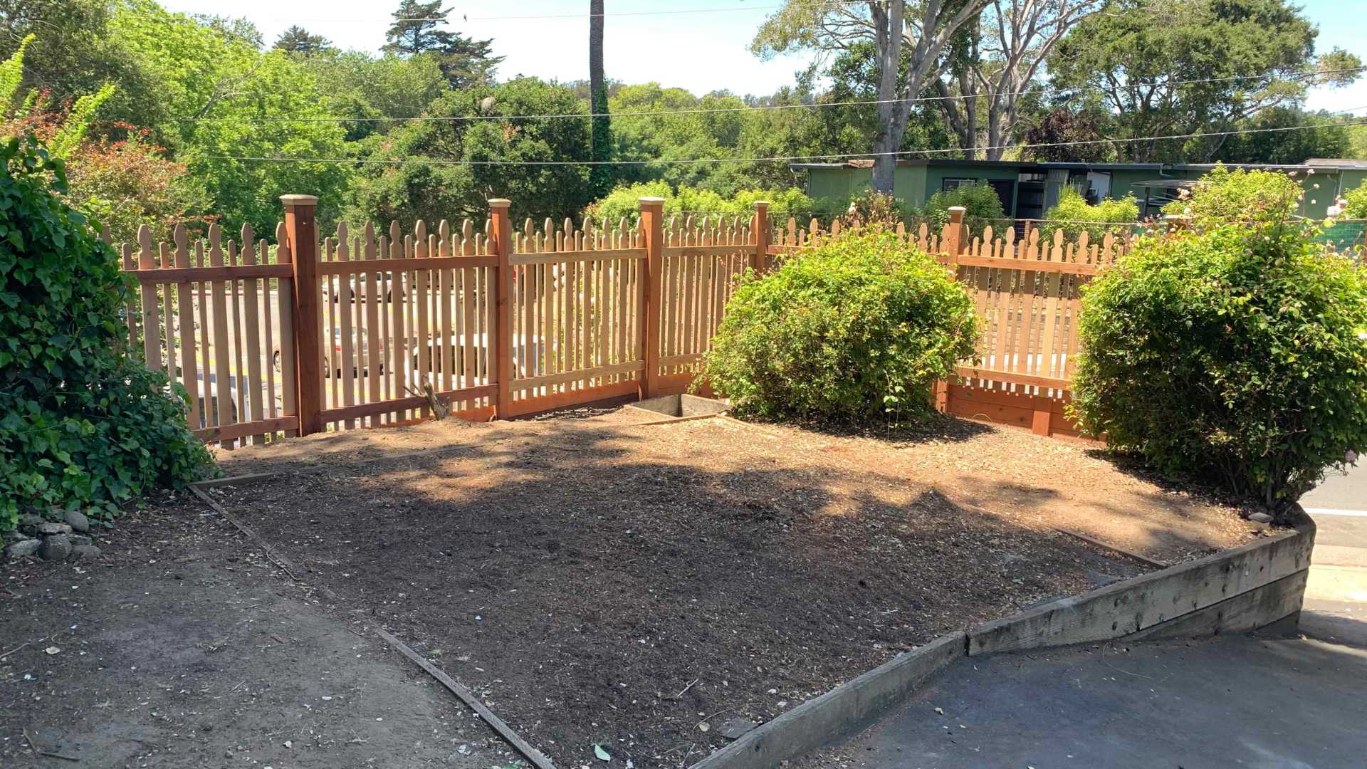 Wood picket fence enclosing small yard with green bushes and trees