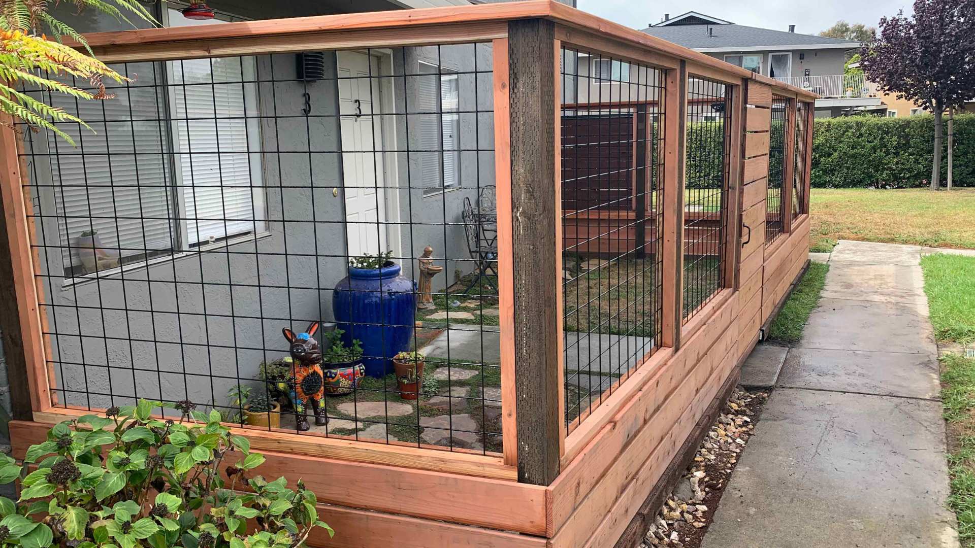 Wood fence with horizontal planks on the bottom and black wire mesh panels on the top, enclosing a small patio area with a blue vase.